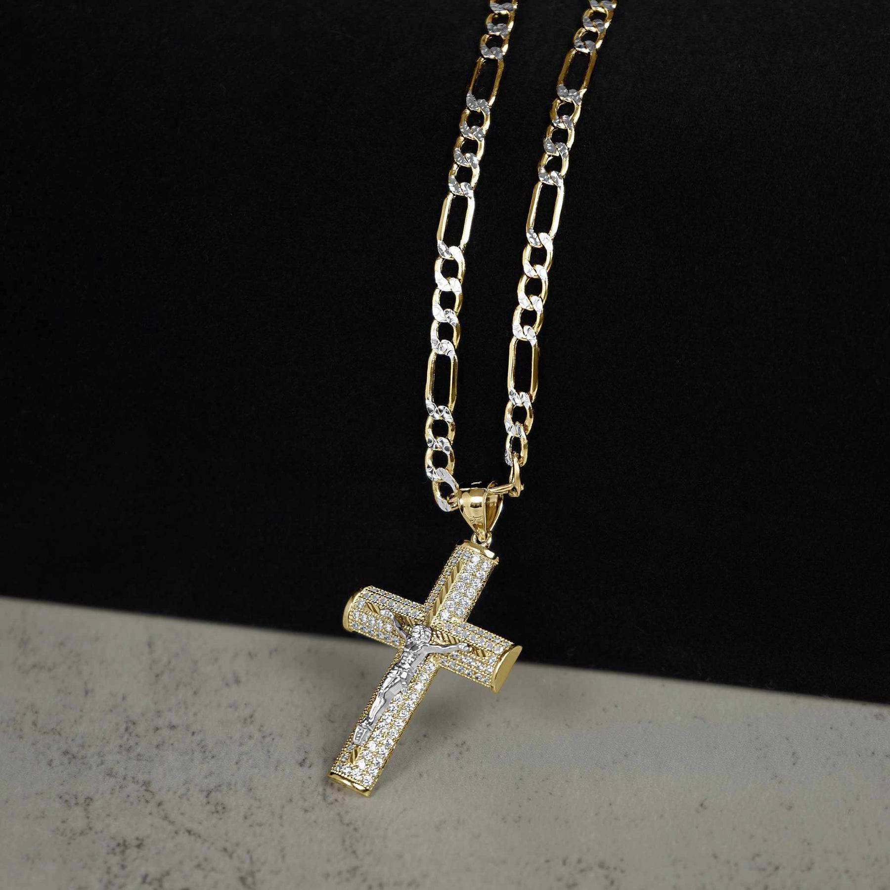 Gold crucifix cross pendant paved with cubic zirconia, hanging from a gold Figaro chain.