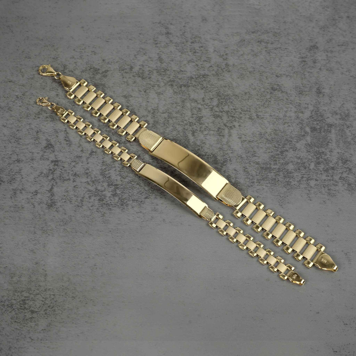 Pair of 10k yellow gold hollow Rollie style ID chain bracelets displayed diagonally on a textured gray background.