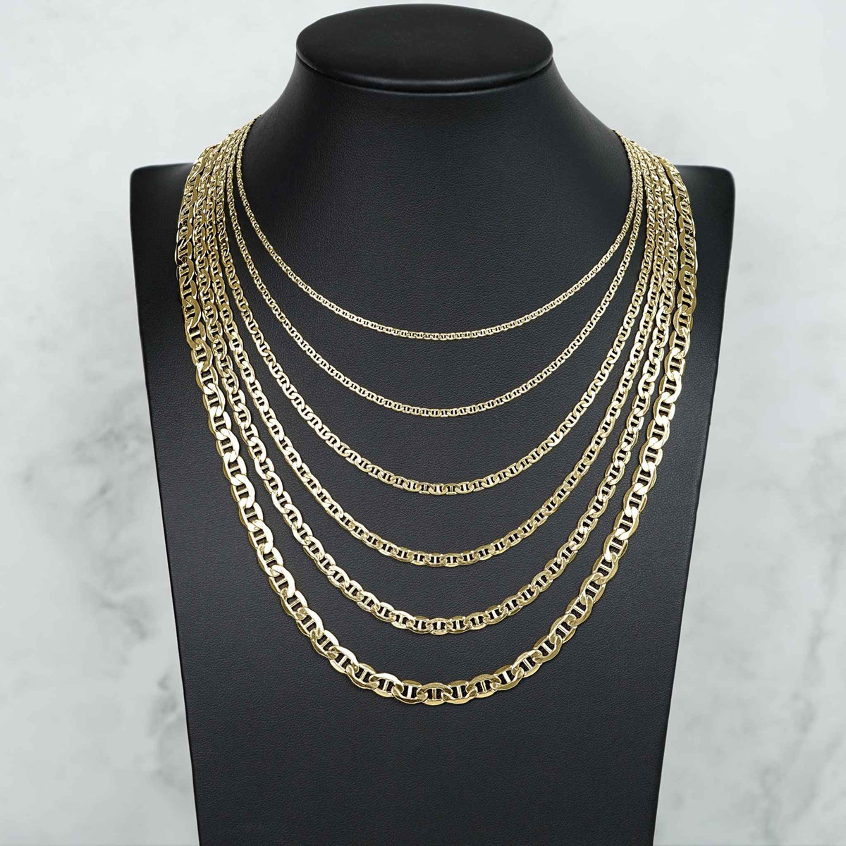 Seven 10k gold mariner chains of varying widths layered on a black jewelry bust.