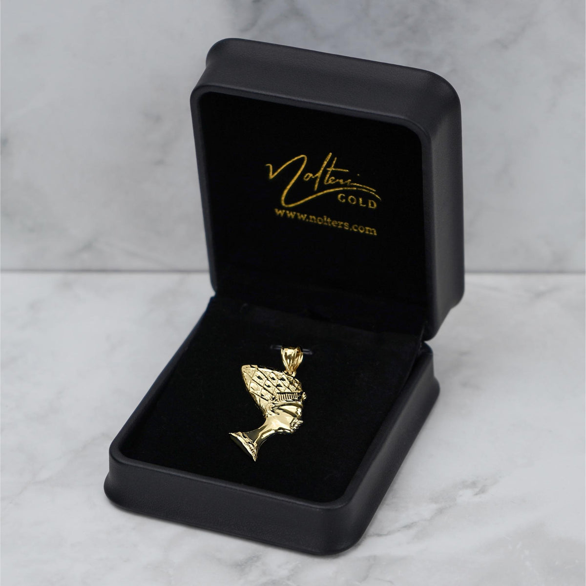 Gold Nefertiti pendant displayed in an open black jewelry box on a marble surface.