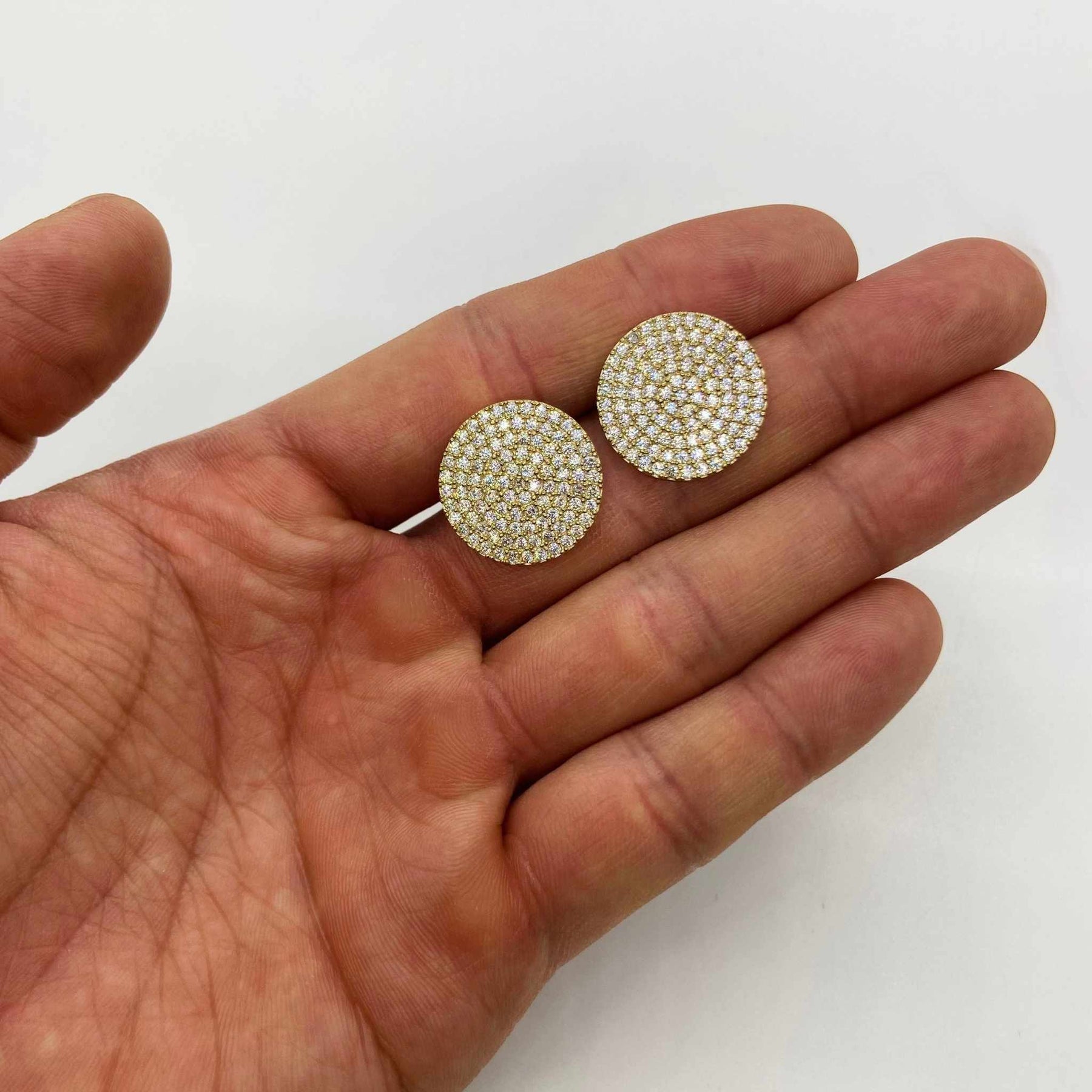 Close-up of a hand holding two large, sparkling gold round CZ stud earrings.
