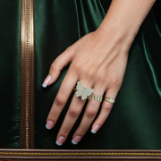 Hand wearing a gold butterfly ring with cubic zirconia against a green background.