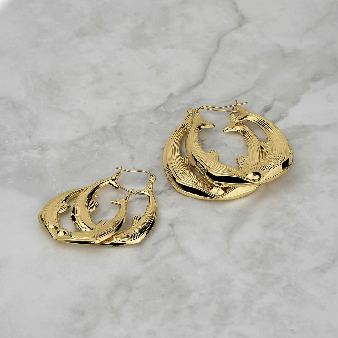 Two sizes of 10k gold kissing dolphin hoop earrings on a light marble surface.