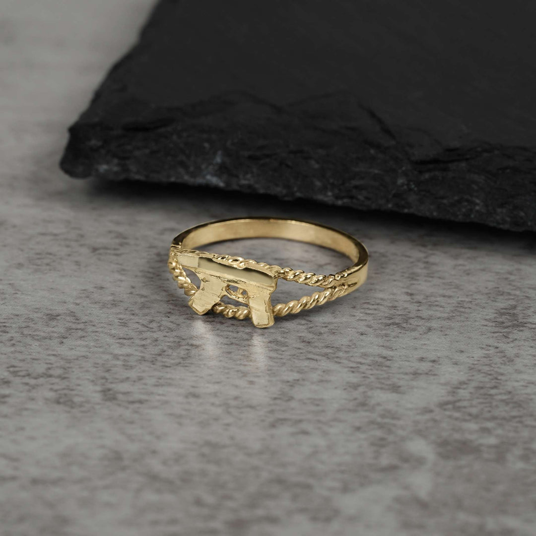 10k gold mini Uzi gun ring with a rope-textured band on gray stone.