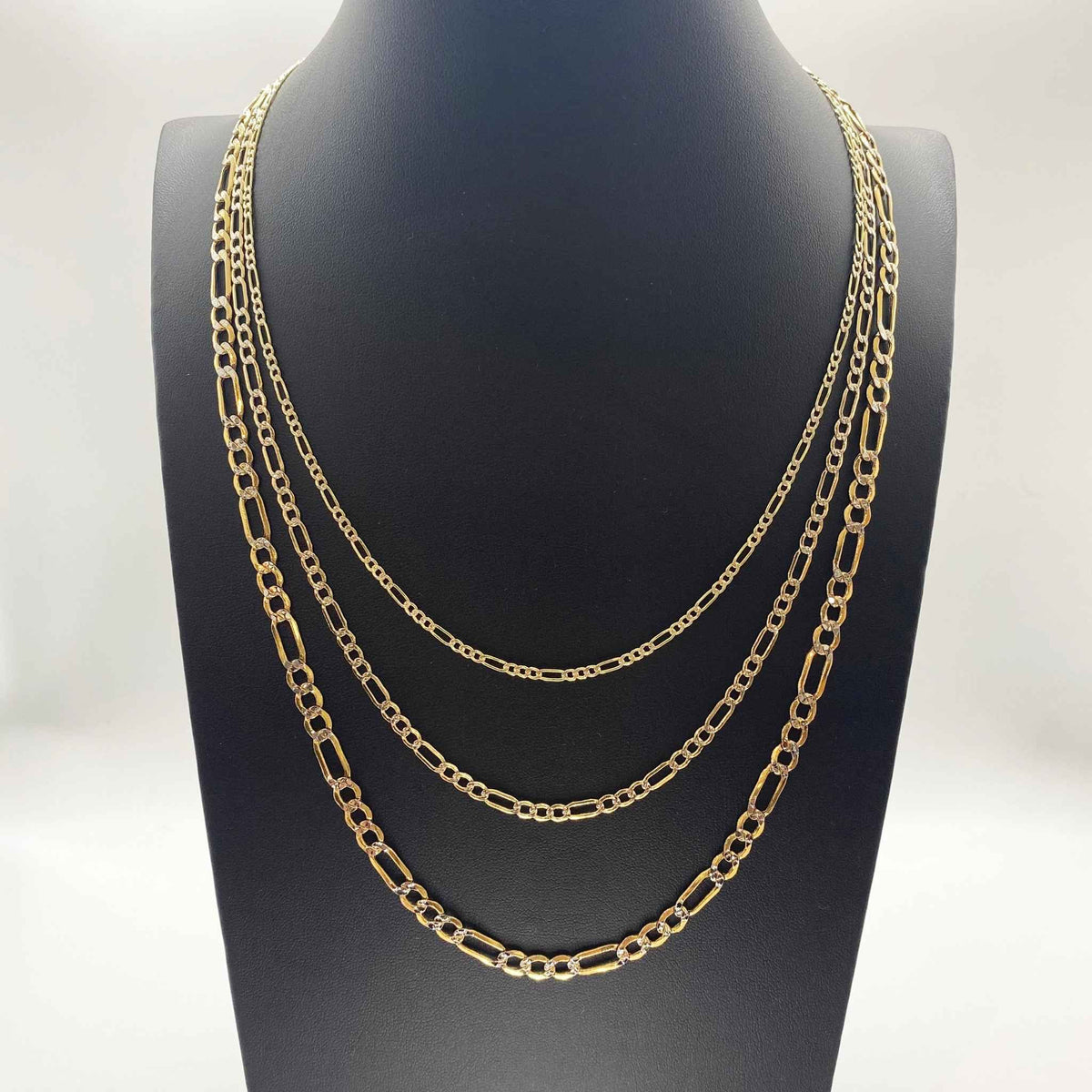 Three gold Figaro chain necklaces of varying widths layered on a black jewelry bust.