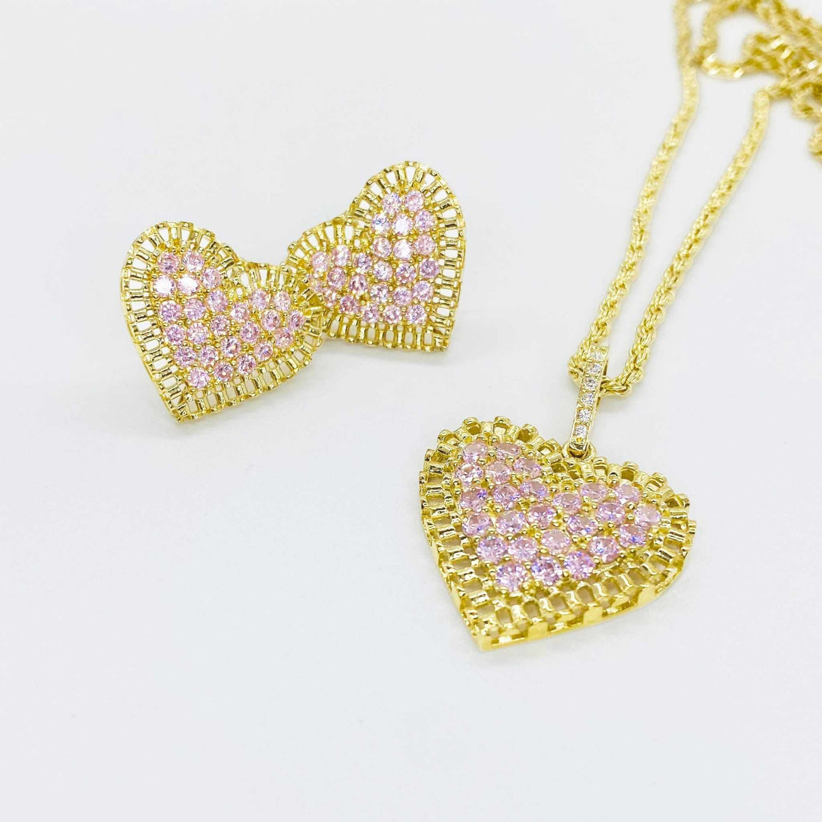 Gold heart pendant necklace and matching stud earrings with pink stones on white background.