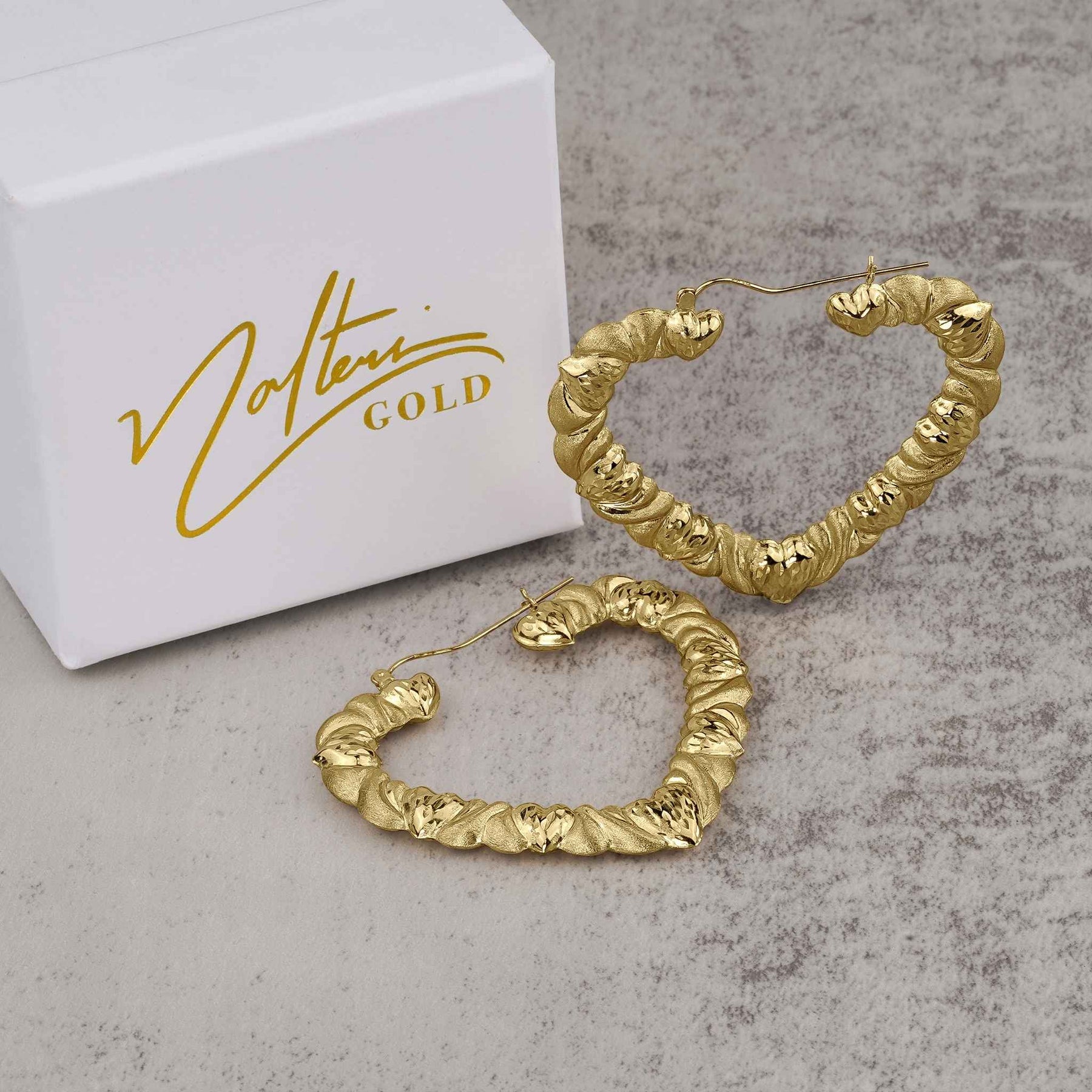 Pair of textured 10k yellow gold twisted heart-shaped hoop earrings on a gray surface.