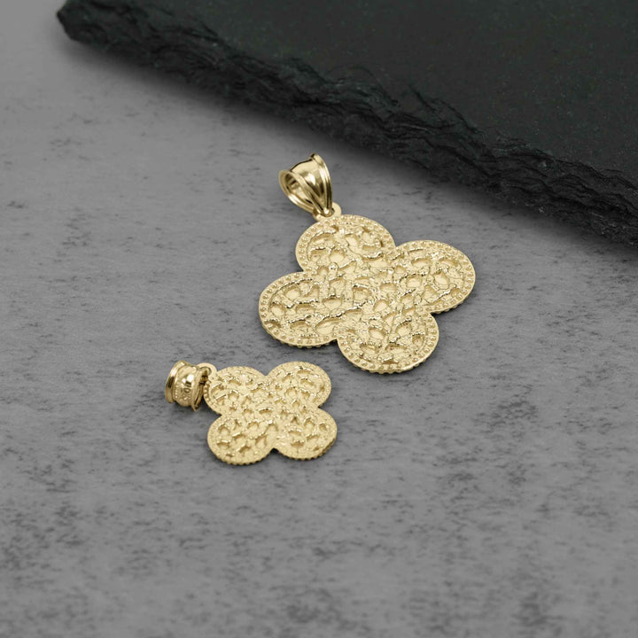 A gold-toned textured clover pendant and matching stud earring displayed on a gray stone surface with a dark slate prop.