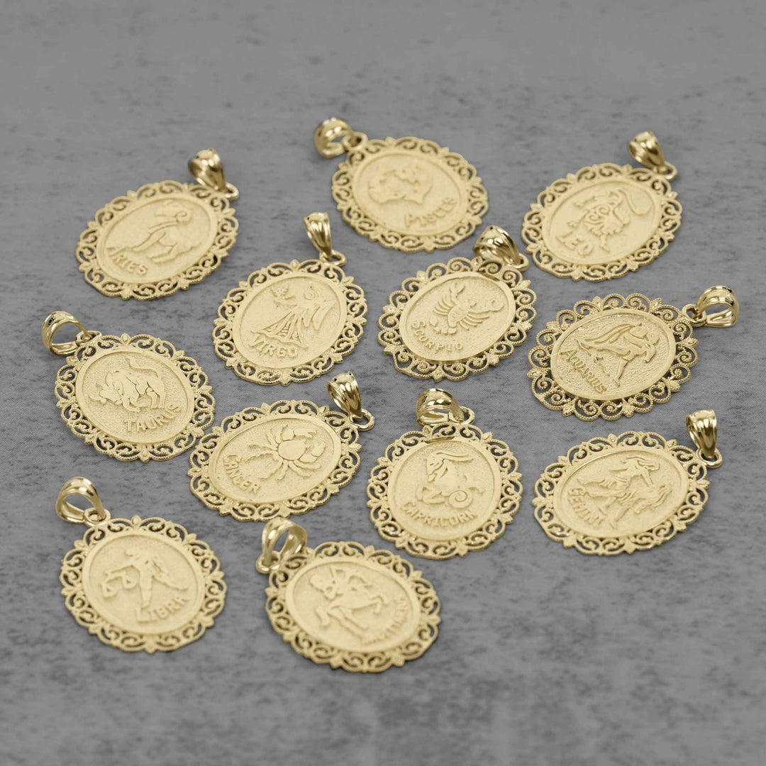 Ten solid 10k yellow gold zodiac pendant charms with ornate filigree borders and raised symbols, arranged on a textured gray background.