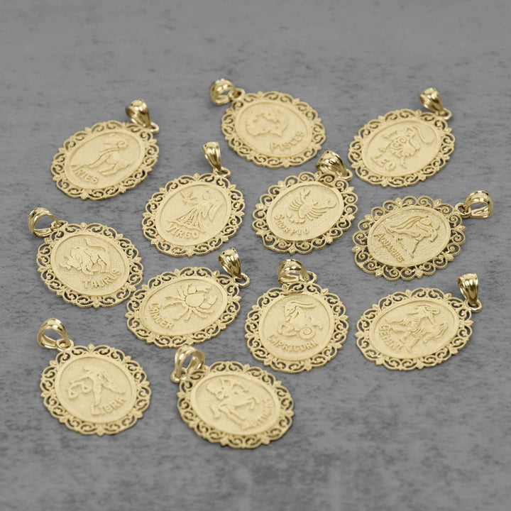 Ten solid 10k yellow gold zodiac pendant charms with ornate filigree borders and raised symbols, arranged on a textured gray background.