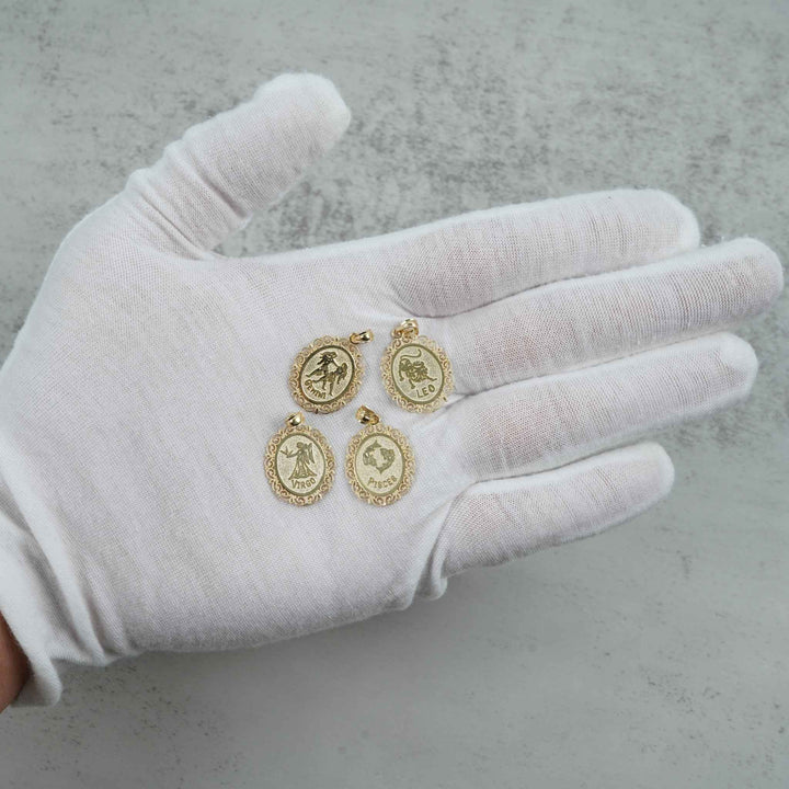 Detailed shot of Aquarius, Scorpio, Leo, and Libra gold zodiac sign pendants held on a white-gloved palm over a neutral surface.