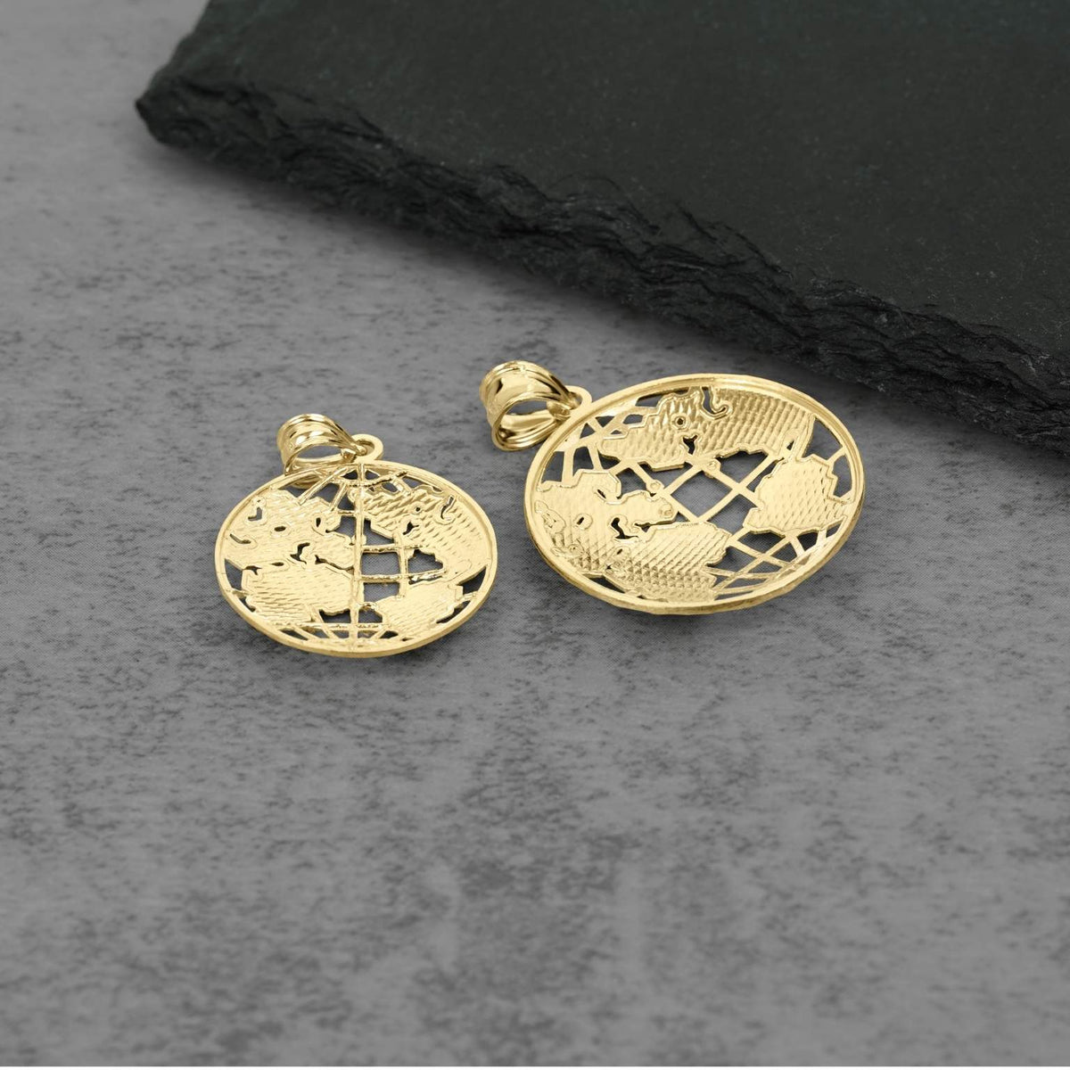 Two gold oval pendant charms with an intricate openwork globe or map design, displayed on a textured gray surface with a black slate prop.