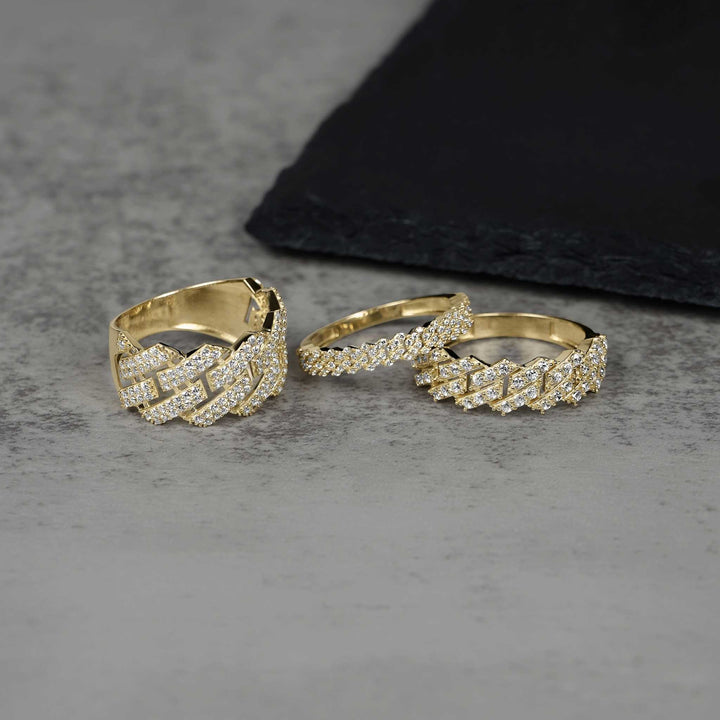 Three iced-out gold Cuban link rings displayed on a gray stone surface.