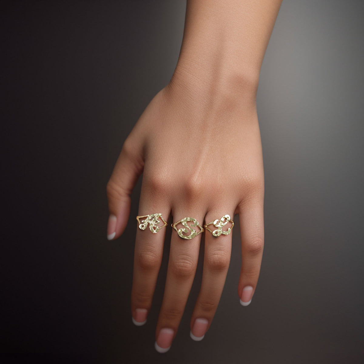 Hand wearing three 10k yellow gold vintage nugget initial rings on a dark background.