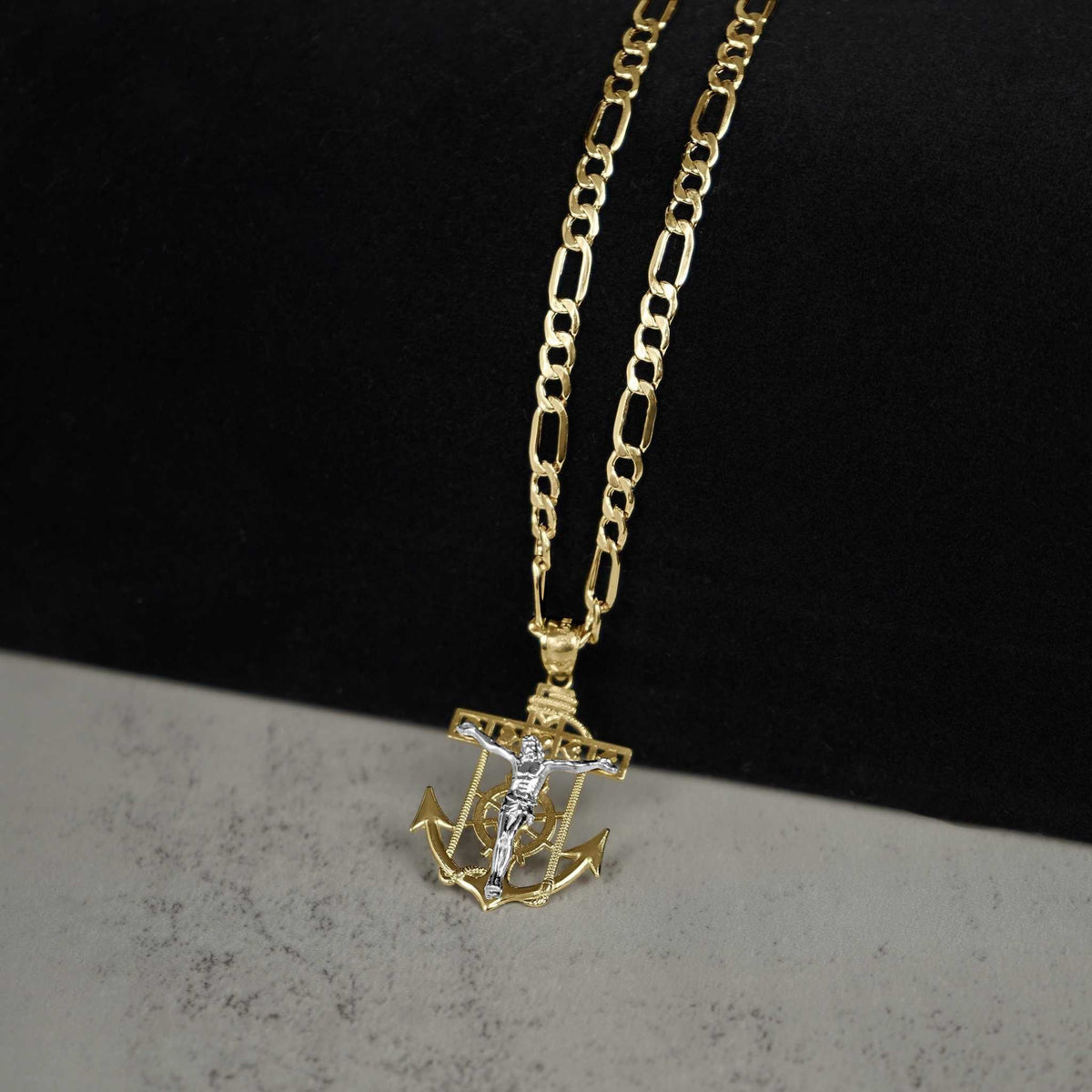 Angled view of a 14k two-tone gold crucifix anchor pendant necklace on a two-tone surface.