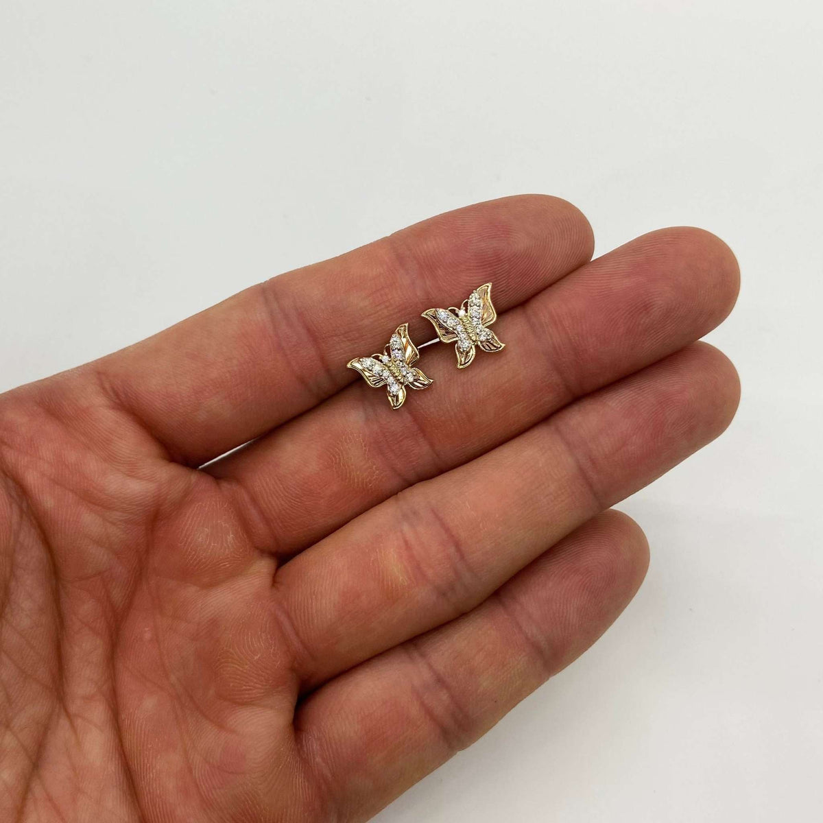 Pair of gold butterfly stud earrings encrusted with clear cubic zirconia held on an open palm.
