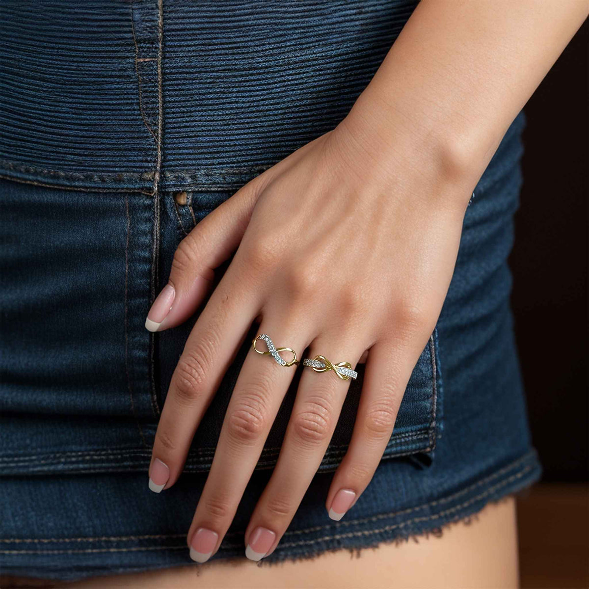 A hand wearing two gold infinity rings accented with cubic zirconia against denim.
