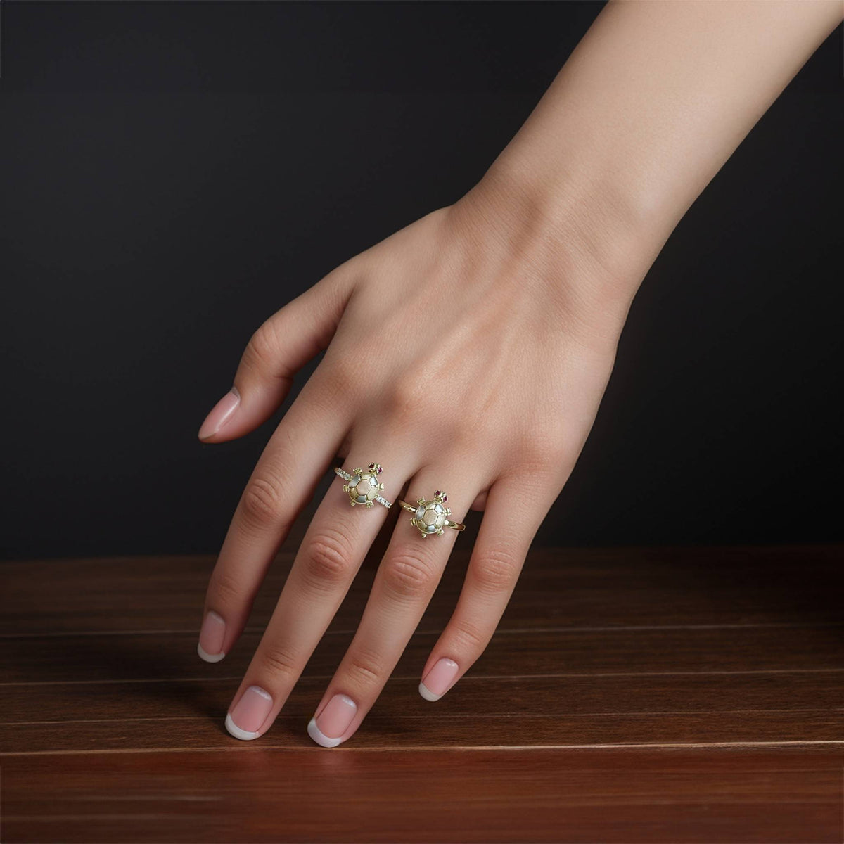 A woman’s hand wearing two tri-color gold turtle rings on a wooden surface.