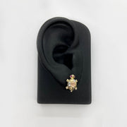 Gold turtle-shaped earring on a black ear model with a white background.