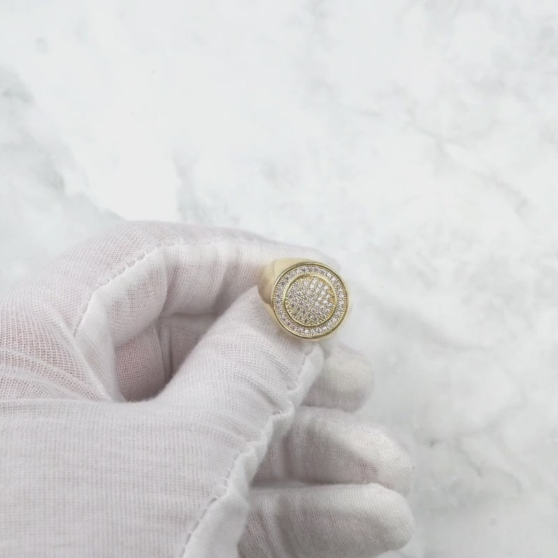 Gloved hand holding and rotating a sparkling gold iced-out round signet ring.