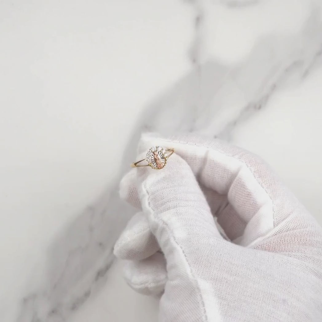 Gloved hand holding a gold Virgin Mary CZ ring over a marble background.