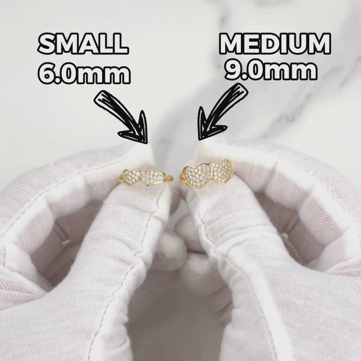 Gloved hands holding small and medium gold dual-heart cubic zirconia rings.