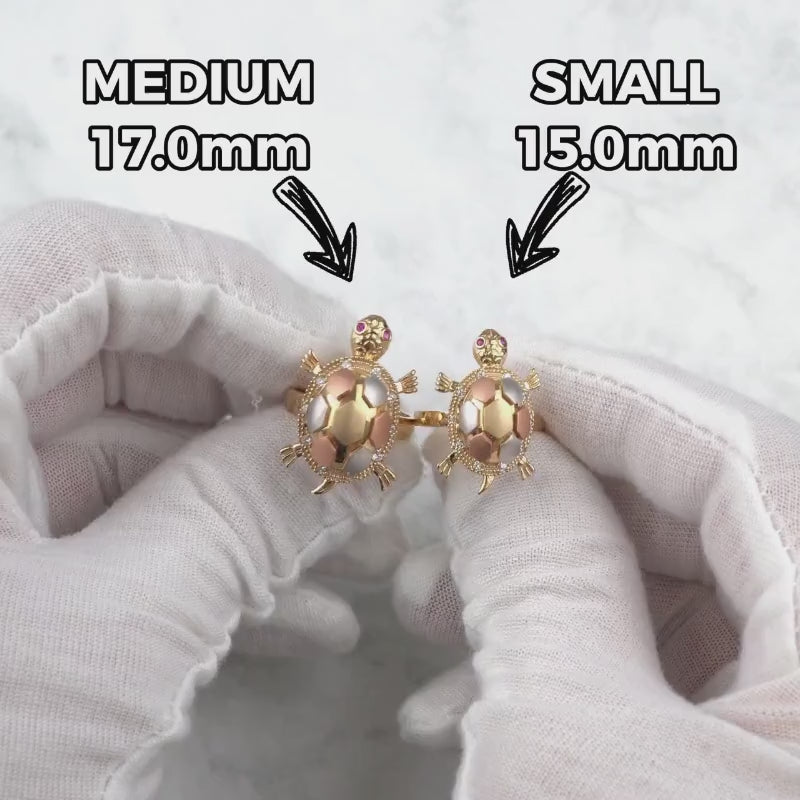 Hands wearing white gloves holding medium and small gold turtle locket rings for size comparison.
